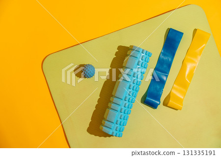 Flat lay on yoga mat with foam roller blue and yellow bands and massage ball over yellow backdrop active recovery 131335191
