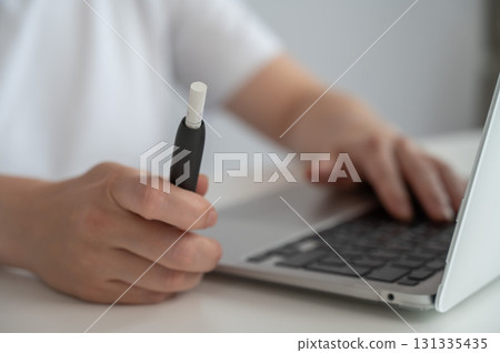 A woman smokes an electronic cigarette while working on a laptop. Tobacco heating system. A woman smokes an electronic cigarette while working on a laptop. Tobacco heating system. 131335435