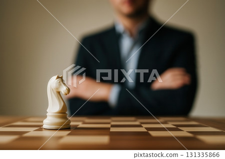 A white chess knight on a wooden chessboard with a blurred businessman in a suit in the background 131335866