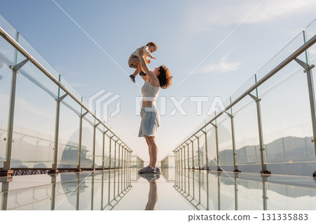 Caucasian woman holds her little son in her arms while standing on a glass bridge in the mountains.  131335883