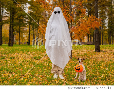 Woman in white sheet and jack russell terrier dog holding jack o latrine in autumn forest. Halloween costume.  131336014