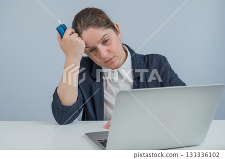 Tired businesswoman smoking an electronic cigarette while working on a laptop. Tobacco heating system. 131336102