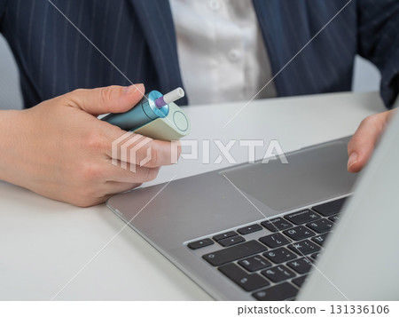 Business woman smoking an electronic cigarette while working on a laptop. Tobacco heating system. Business woman smoking an electronic cigarette while working on a laptop. Tobacco heating system. 131336106