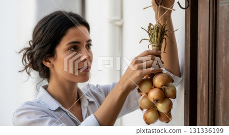 Woman hanging a bunch of fresh yellow onions in a rustic setting. Storing harvested vegetables for cooking. Natural food and healthy lifestyle concept Woman hanging a bunch of fresh yellow onions in a rustic setting. Storing harvested vegetables for cooking. Natural food and healthy lifestyle concept 131336199