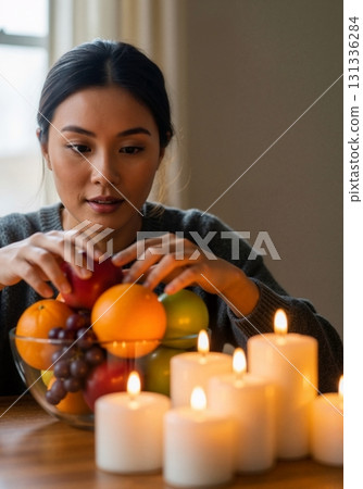 Young Asian woman arranging a fruit bowl by candlelight. Cozy home atmosphere for wellness and relaxation. Healthy lifestyle and self-care concept Young Asian woman arranging a fruit bowl by candlelight. Cozy home atmosphere for wellness and relaxation. Healthy lifestyle and self-care concept 131336284