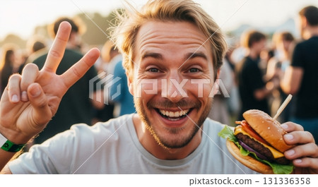 Happy young man eating a burger and showing a peace sign at a summer festival. Portrait of a smiling person enjoying fast food outdoors at sunset Happy young man eating a burger and showing a peace sign at a summer festival. Portrait of a smiling person enjoying fast food outdoors at sunset 131336358