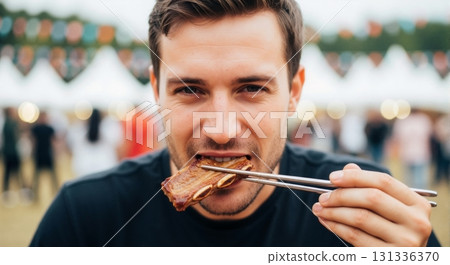Young man eating a barbecue rib with chopsticks at an outdoor food festival. Close-up portrait of a person enjoying street food 131336370