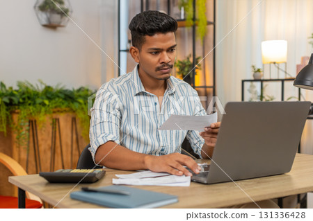Indian man uses laptop and calculator to enter bill numbers compare sums plan budget savings finance 131336428
