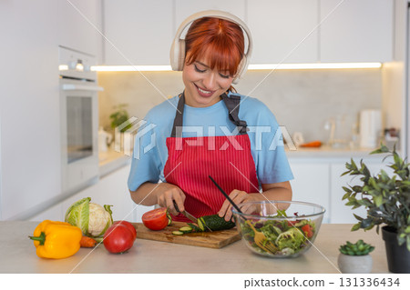 Young woman with headphones plays anti stress music cuts cucumbers and tomatoes for fresh salad prep 131336434