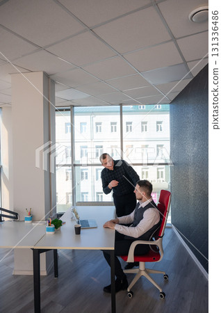 Caucasian man standing at colleague's desk discussing work.  131336486