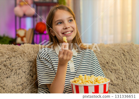 Child girl kid happily eats popcorn while watching cartoon on TV and laughing cheerfully on sofa Child girl kid happily eats popcorn while watching cartoon on TV and laughing cheerfully on sofa 131336651
