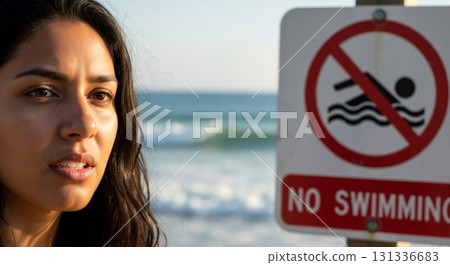 Contemplative woman on the beach with a no swimming warning sign. Close-up portrait focusing on safety and prohibition. Ocean waves in the background 131336683