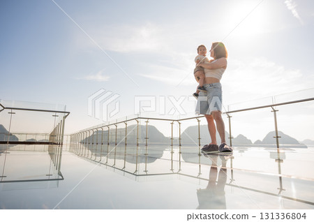 Caucasian woman holds her little son in her arms while standing on a glass bridge in the mountains.  131336804