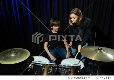 Young caucasian woman teaches a boy to play the drums in the studio on a black background. Music school student 131337116