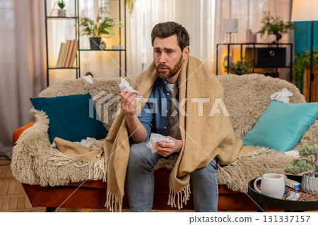 Young man sitting on home sofa, wrapped in a blanket, sneezing, reaching for tissues, feeling sick Young man sitting on home sofa, wrapped in a blanket, sneezing, reaching for tissues, feeling sick 131337157