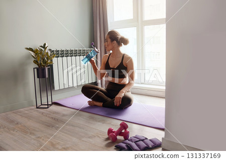 Woman drinking water sitting on exercise mat at home Woman drinking water sitting on exercise mat at home 131337169
