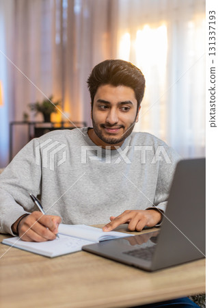 Young Indian man student attending online class through laptop while taking notes at table at home Young Indian man student attending online class through laptop while taking notes at table at home 131337193