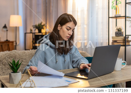 Focused woman freelancer reading documents and using laptop computer at table in living room at home 131337199