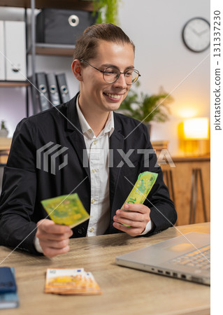 Happy young Caucasian businessman counting money cash and using laptop computer in home office Happy young Caucasian businessman counting money cash and using laptop computer in home office 131337230