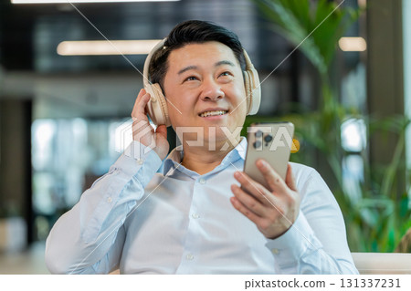 Excited mature businessman in headphones listening energetic music on smartphone in office lobby Excited mature businessman in headphones listening energetic music on smartphone in office lobby 131337231