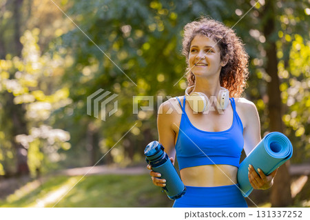 Young happy sportswoman with mat and water bottle in hands is heading to training outdoors in park Young happy sportswoman with mat and water bottle in hands is heading to training outdoors in park 131337252