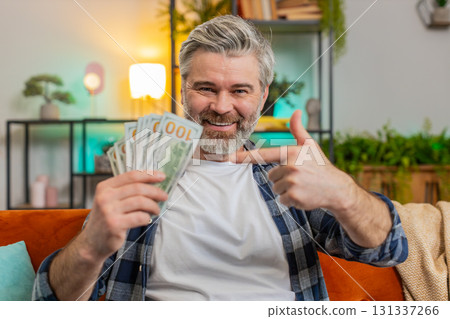 Amazed middle-aged man showing pointing money dollar cash bills clenching fist thumbs up at home Amazed middle-aged man showing pointing money dollar cash bills clenching fist thumbs up at home 131337266