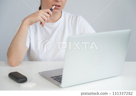A woman smokes an electronic cigarette while working on a laptop. Tobacco heating system. A woman smokes an electronic cigarette while working on a laptop. Tobacco heating system. 131337282