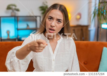 Furious young Caucasian woman sitting on sofa at home, angrily pointing at you through the camera 131337284