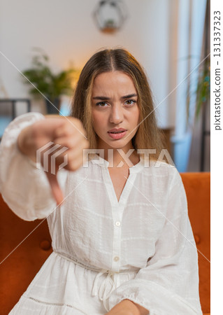 Upset young woman showing thumbs down sign gesture, expressing discontent sitting on sofa at home 131337323