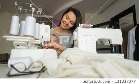 A young woman sews in the kitchen and holds a small child. Mom teaches her little son to sew on a sewing machine. Self-isolation homework A young woman sews in the kitchen and holds a small child. Mom teaches her little son to sew on a sewing machine. Self-isolation homework 131337377