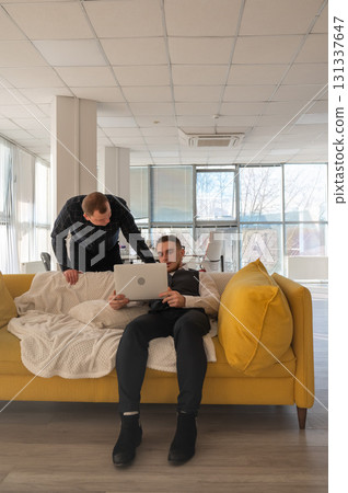 Two Caucasian men discussing work on a laptop while sitting on a sofa. Modern office.  131337647