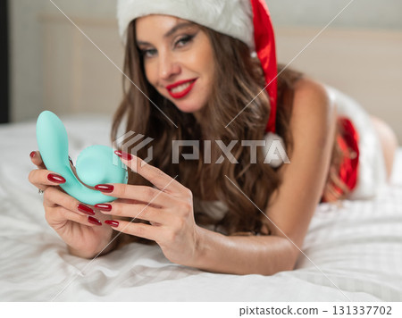 Caucasian woman in Santa Claus costume lying on bed holding curved sex toy.  131337702