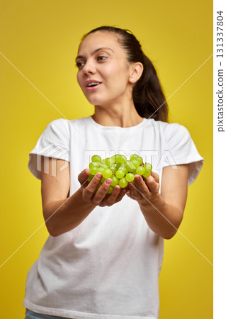portrait of attractive Caucasian smiling woman holds grapes isolated on yellow studio background, focus on grapes 131337804