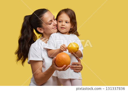 Cheerful little child girl with mother holding lemon and grapefruit on yellow background. happy family having fun together Cheerful little child girl with mother holding lemon and grapefruit on yellow background. happy family having fun together 131337836