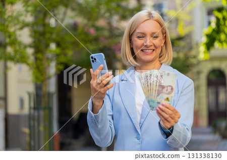Caucasian mature businesswoman on city street showing earnings cash money with smartphone in hands Caucasian mature businesswoman on city street showing earnings cash money with smartphone in hands 131338130