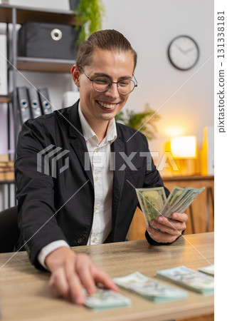 Successful young businessman guy in formal suit sitting counting money at table in home office 131338181
