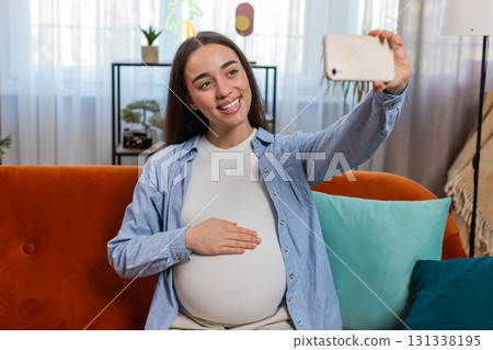 Happy pregnant woman sits on sofa taking selfie using smartphone touching belly enjoying motherhood 131338195