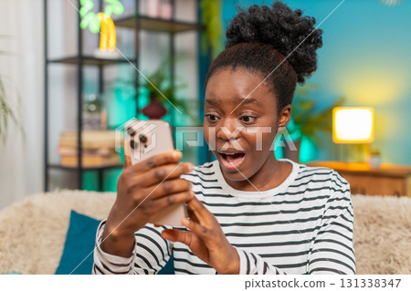 African American woman surprised smartphone wide eyes expression unexpected message indoors sofa African American woman surprised smartphone wide eyes expression unexpected message indoors sofa 131338347