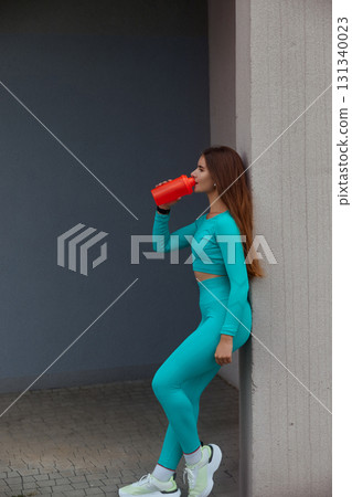 Fit woman in turquoise activewear drinks from a red water bottle while leaning against a wall; showcasing a healthy lifestyle and fitness routine. Workout concept Fit woman in turquoise activewear drinks from a red water bottle while leaning against a wall; showcasing a healthy lifestyle and fitness routine. Workout concept 131340023