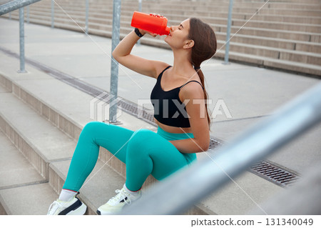 Young Woman Drinks Water While Resting on Stairs After Workout in Urban Environment During Daylight 131340049