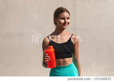 Fit young woman in a black sports bra holds a bright orange shaker bottle, standing confidently against a textured wall, embodying a healthy lifestyle and fitness motivation. fit, sport, workout 131340072