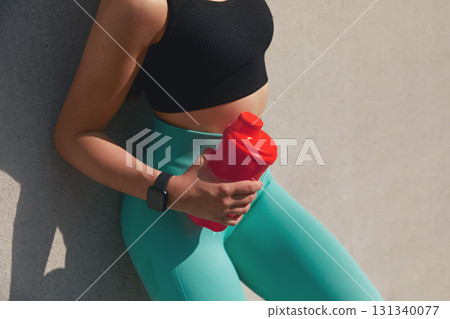 Fit woman in black sports bra and turquoise leggings holds a red water bottle while leaning against a textured wall, embodying a healthy lifestyle and fitness motivation. 131340077