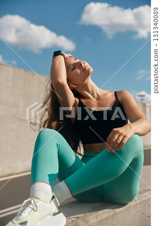 Fit woman in athletic wear sitting on a concrete barrier, enjoying the sun with a relaxed expression, showcasing a sporty lifestyle and the joy of outdoor fitness activities. Fit woman in athletic wear sitting on a concrete barrier, enjoying the sun with a relaxed expression, showcasing a sporty lifestyle and the joy of outdoor fitness activities. 131340089
