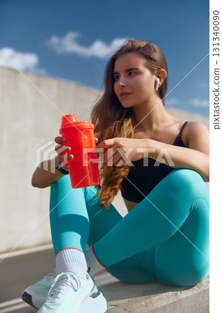 Fit woman with long hair sitting on a ledge, holding a bright red shaker bottle, wearing stylish activewear, enjoying a moment of relaxation after an intense workout session outdoors. 131340090