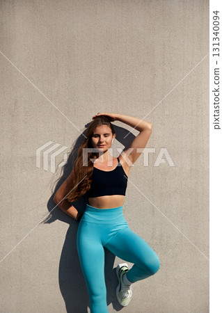 Fit woman in athletic wear poses confidently against a textured wall, showcasing strength and determination, embodying the spirit of fitness and wellness in a vibrant workout environment. 131340094
