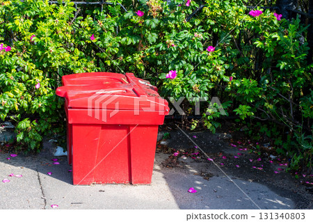 A red trash can is currently sitting on the sidewalk near a bush A red trash can is currently sitting on the sidewalk near a bush 131340803