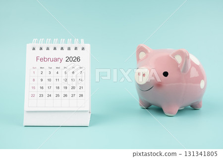 February 2026 Desk calendar and pink piggy bank on blue background. February 2026 Desk calendar and pink piggy bank on blue background. 131341805