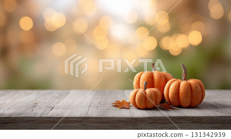 Three pumpkins of varying sizes sit on a weathered wooden table alongside fallen autumn leaves, creating a warm and inviting autumn scene with soft bokeh lights in the background 131342939