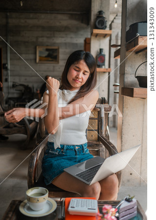 A young woman takes a break to stretch while working on her laptop in a stylish, rustic cafe setting. 131343009