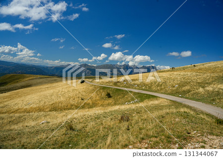 Scenic road winding through golden hills with mountains in the background under a bright blue sky with white clouds. Perfect for travel, landscape, and nature imagery 131344067